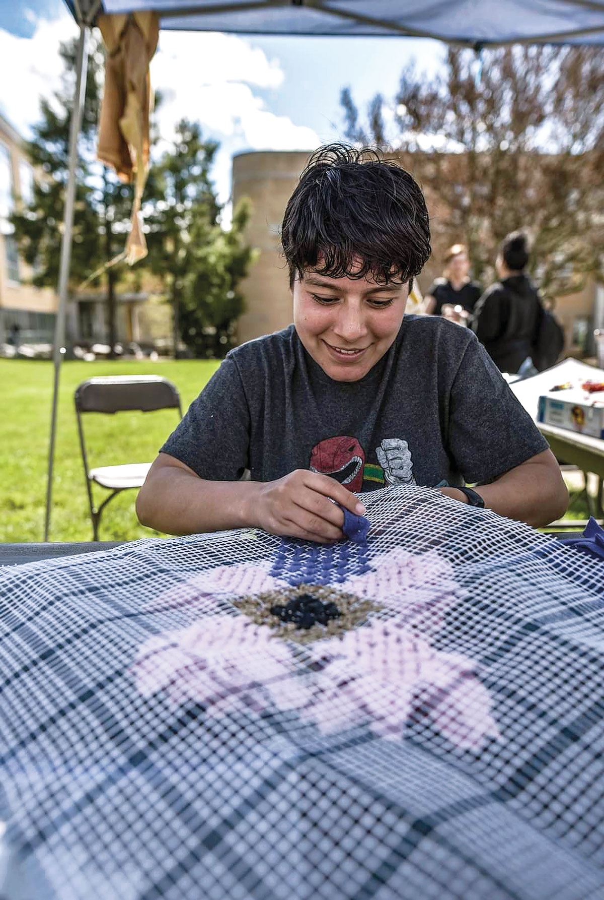 boy sits at table weaving blue string into pattern on cloth